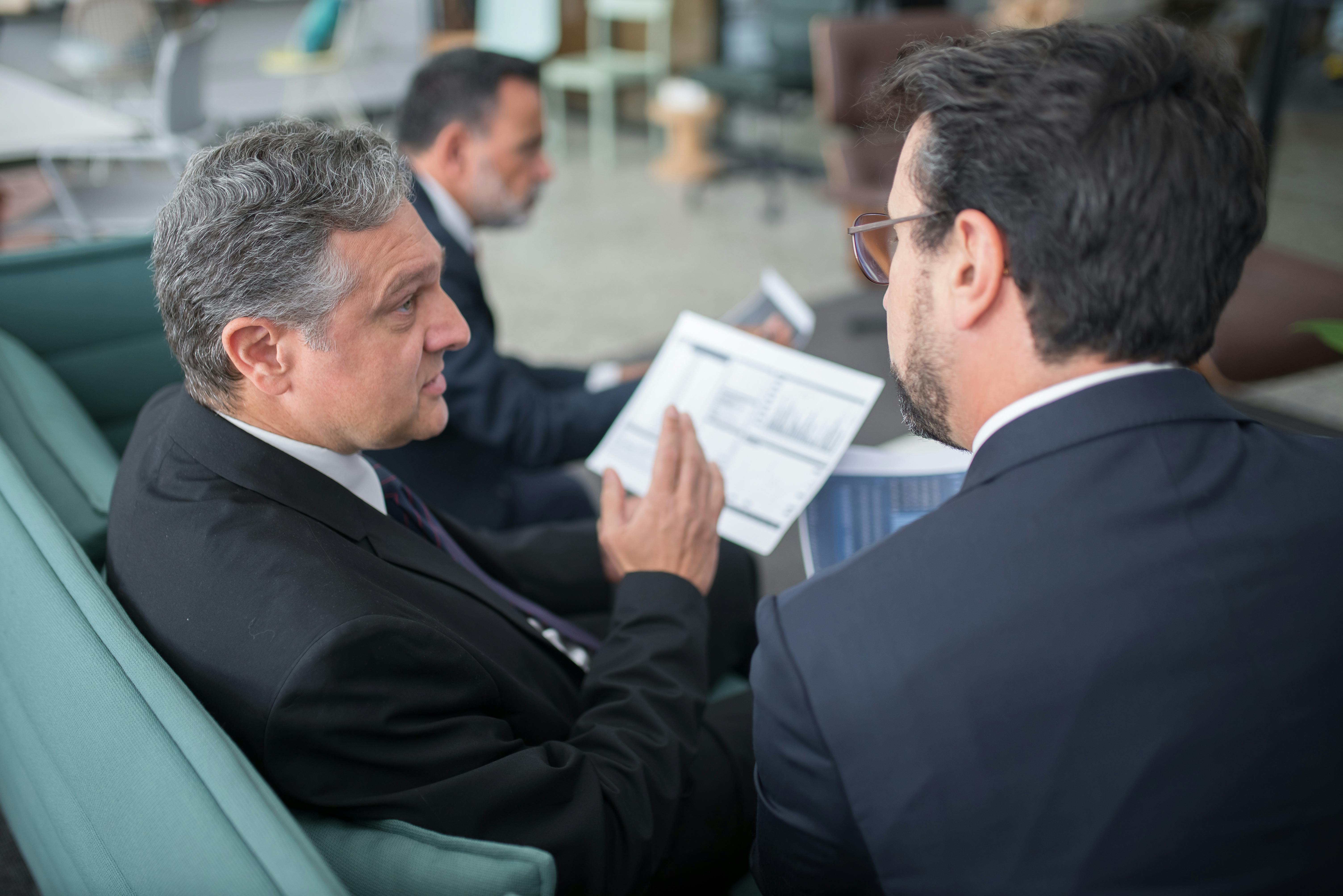 Two businessmen in suits discussing financial reports while seated on a couch in a modern office setting.